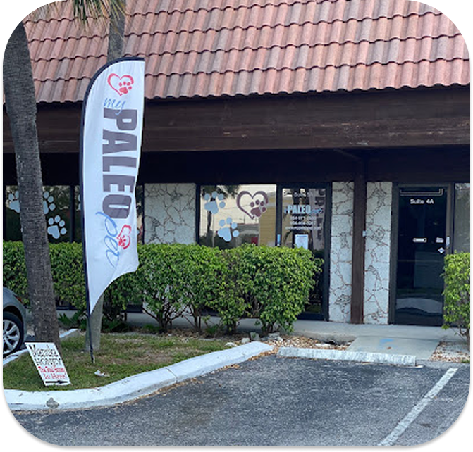 Exterior view of the My Paleo Pet store with a tiled roof, stone facade, and a vertical banner featuring heart and paw print designs, located at Suite 104 with green shrubs and a parking lot.