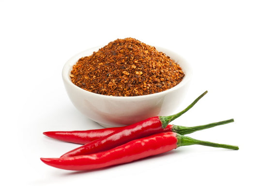 A white bowl filled with red cayenne powder, alongside three whole red chili peppers on a white background.