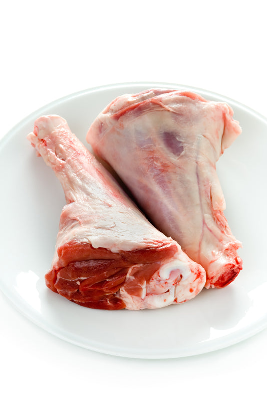 Two raw lamb shank bones on a white plate against a white background.