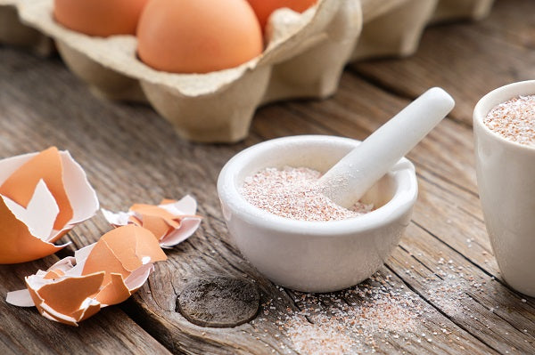 Ground eggshell powder in a white bowl with a spoon, next to a carton of brown eggs and eggshells on a wooden surface.