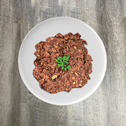 A bowl of ground natural raw chicken mix, presumably the product mentioned, garnished with a leaf of parsley on a wooden surface.