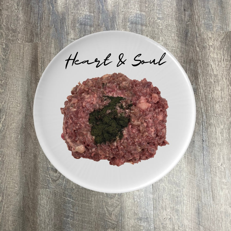 A plate of ground pork meat with visible pieces of herbs or spices, placed on a wooden surface, with the words 'Heart & Soul' written above it.