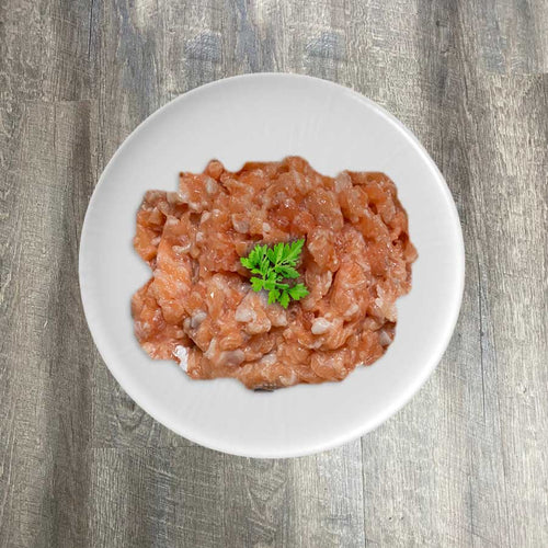 A bowl of raw ground salmon on a plate, with a sprig of parsley for garnish, on a wooden surface.