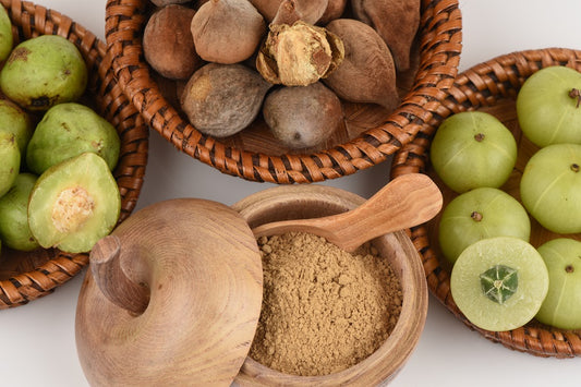 The image shows baskets of fresh amla fruits, along with a wooden bowl of Triphala powder and a spoon, against a white background.
